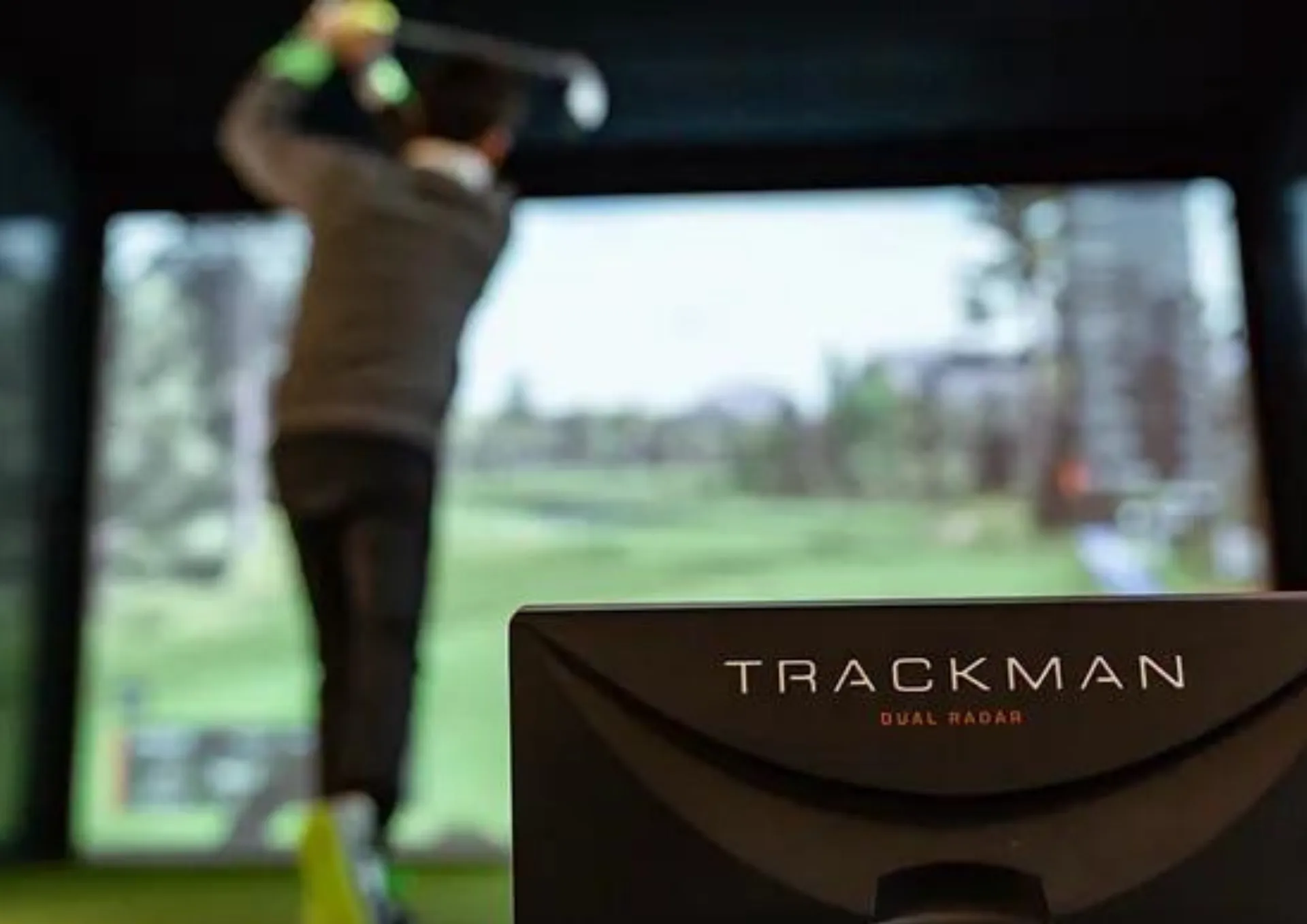 Golfeur en plein swing devant l'écran du simulateur, radar Trackman au premier plan